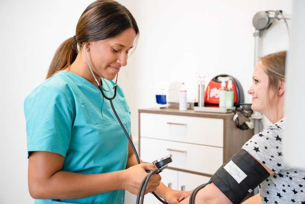 Das Bild zeigt eine freundliche medizinische Fachangestellte, die eine Blutdruckmessung an einer Patientin macht
